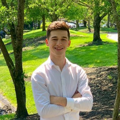 Justin Biller, smiling, arms crossed, standing outdoors among trees in a white shirt